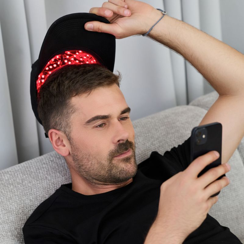 man_wearing_red_light_therapy_cap_and_holding_up_while_looking_at_phone